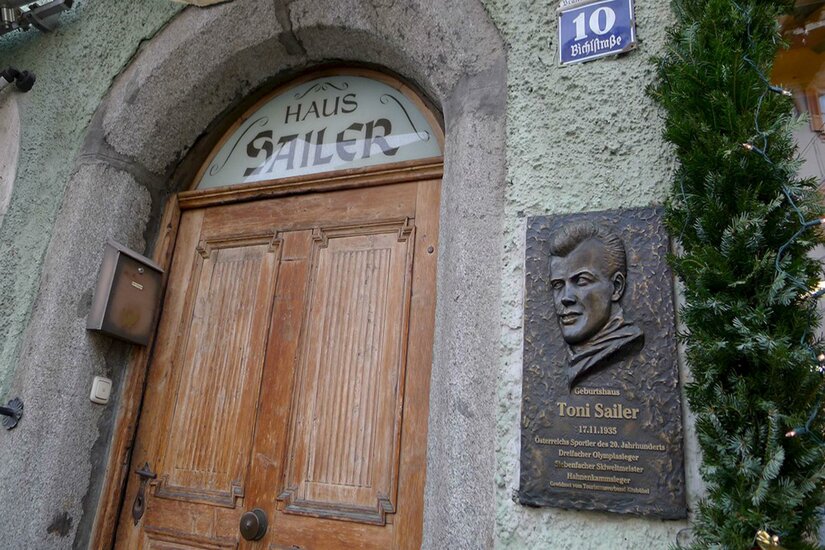 Eingang Haus Sailer Holztür unter Steinbogen mit der Aufschrift Haus Sailer und einer Gedenktafel für Toni Sailer daneben.