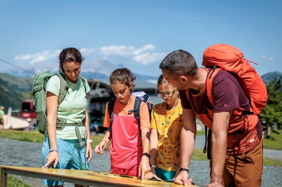 Eine vierköpfige Familie mit Wanderrucksäcken betrachtet gemeinsam eine Informationstafel im Freien.