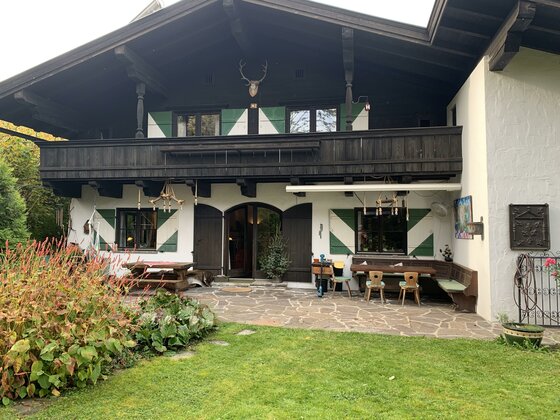 Garten1 Ein traditionelles Haus im ländlichen Stil mit einem großen Balkon und bunten Fensterläden. Der Garten ist grün und einladend, mit einem gemütlichen Sitzbereich.