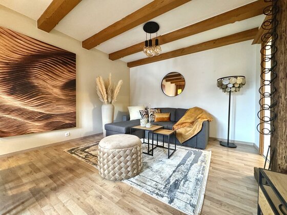 Ein modernes Wohnzimmer mit einer bequemen Sofa-Ecke und einer eleganten Lampe. Die Wände sind hell, und es gibt ein großes Wandbild sowie einen runden Tisch im Zentrum des Raumes.