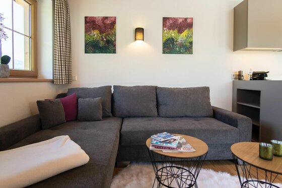 Wohnbereich im Hof Zenzern Graues Ecksofa mit Kissen vor einer weißen Wand mit zwei Bildern und einer Wandlampe.