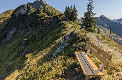 Holzbank an einem schmalen Wanderpfad auf einem grasbewachsenen Bergrücken mit Blick auf Gipfel.
