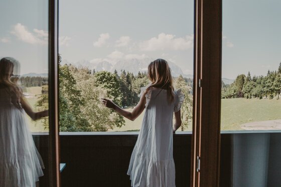 Blick vom Hotel Seebichl Frau in weißem Kleid blickt von einem Balkon auf eine Wald- und Berglandschaft.