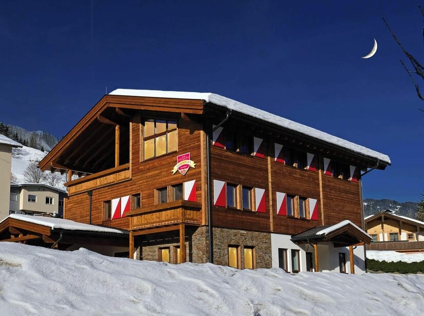 Dreistöckiges Holzhaus mit rot-weißen Fensterläden im Schnee unter blauem Himmel mit Mondsichel.