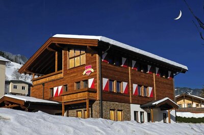 Dorfstube Jochberg Winteransicht Dreistöckiges Holzhaus mit rot-weißen Fensterläden im Schnee unter blauem Himmel mit Mondsichel.