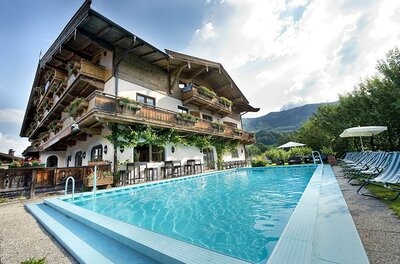 Außenpool vor einem mehrstöckigen alpinen Hotelgebäude mit Holzbalkonen und Liegestühlen.
