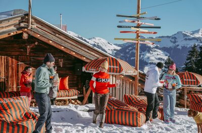 Après-Ski bei der Schilala Personen vor einer Holzhütte im Schnee mit Sitzsäcken und einem Wegweiser aus Skiern.