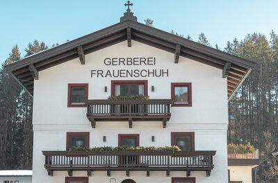 WEB_HansFrauenschuh_-66 Ein traditionelles, weißes Gebäude mit einem großen Balkon und Holzverzierungen. Es trägt den Namen "Gerberei Frauenschuh" und befindet sich in einer ländlichen Umgebung.