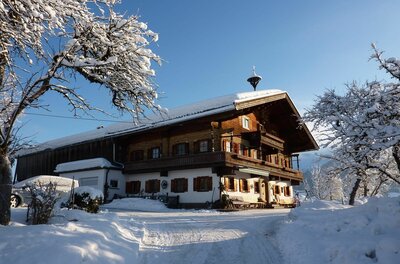Hörpfing im Winter Verschneiter Bauernhof aus Holz und Stein unter blauem Himmel mit schneebedeckten Bäumen im Vordergrund.
