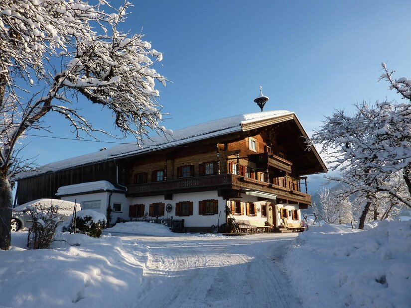 Hörpfing im Winter Verschneiter Bauernhof aus Holz und Stein unter blauem Himmel mit schneebedeckten Bäumen im Vordergrund.