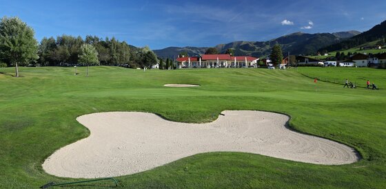 Golf Club Urslautal Golfplatz mit Sandbunker im Vordergrund, grüner Rasenfläche und Clubhaus vor Bergkulisse.
