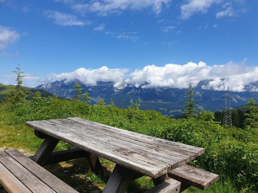 Rastplatz am Rauhen Kopf Holzpicknicktisch auf einer Wiese vor einem Bergpanorama mit Wolken.