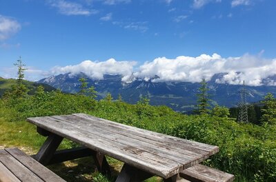 Holzpicknicktisch auf einer Wiese vor einem Bergpanorama mit Wolken.