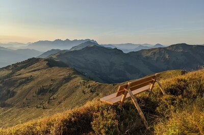 Holzbank auf einem grasbewachsenen Berggipfel mit Blick auf eine weite Gebirgskette bei Sonnenuntergang.