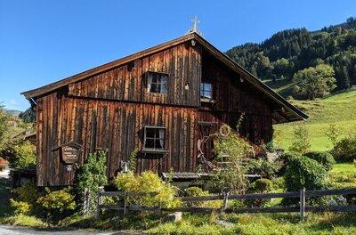 Altes Holzhaus mit dunkler Fassade und kleinem Gipfelkreuz vor bewaldetem Hang.