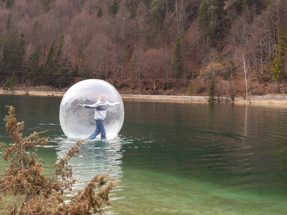 Fun Ball Eine Person steht in einer transparenten Ballon-Kugel auf einem ruhigen See. Im Hintergrund sind Bäume und sanfte Hügel zu sehen.