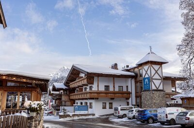 Außenansicht des Dorfresort Kitzbühel mit schneebedeckten Dächern und Parkplatz vor dem Gebäude.