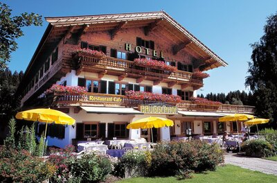 Bruggerhof im Sommer Traditionelles Holzhotel mit Restaurantterrasse, gelben Sonnenschirmen und Blumenschmuck unter blauem Himmel.