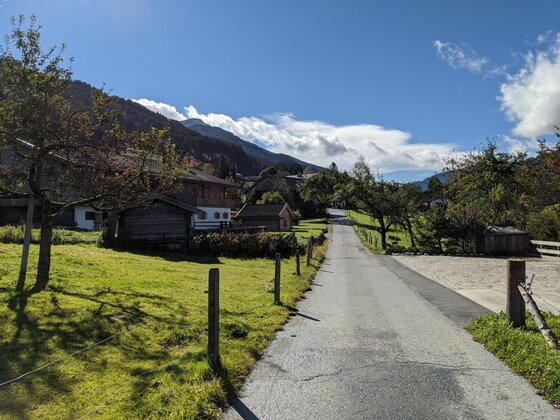 Museumsweg Jochberg Asphaltierter Weg führt an Holzhütten und grünen Wiesen in Jochberg vorbei.