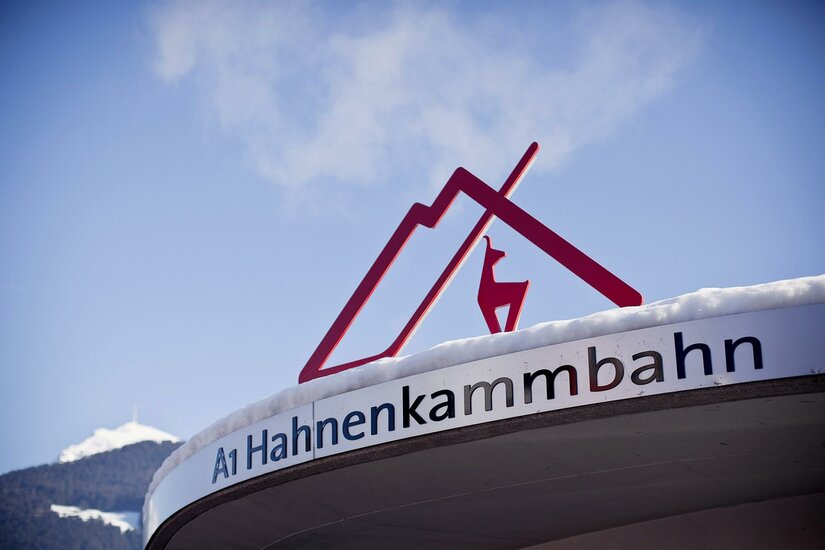 Hahnenkammbahn Ein Schild der Hahnenkammbahn mit einem stilisierten Dach und einem Reh. Der Schreibstil ist modern und ansprechend, vor einer bergigen Kulisse.