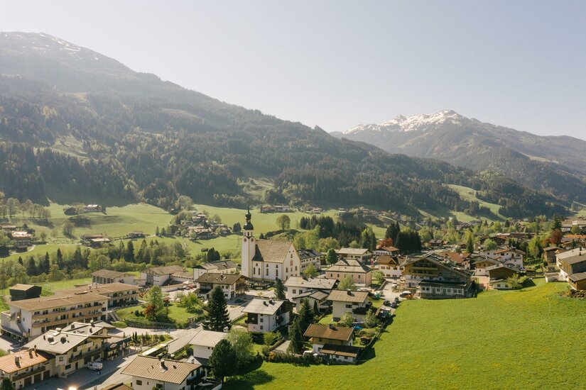 Luftaufnahme eines Alpendorfes mit Kirche vor bewaldeten Bergen und schneebedeckten Gipfeln.