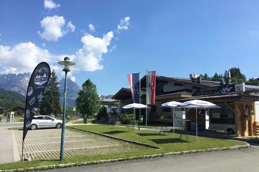 Außenansicht des Kulturhauses mit Imbissstand, Sonnenschirmen und Parkplatz vor Bergpanorama.