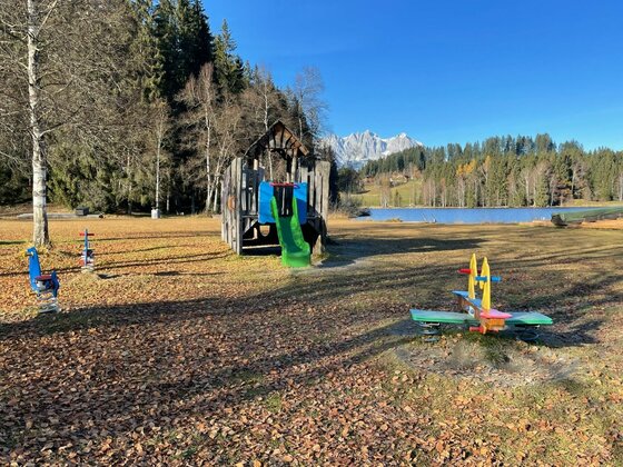 Spielplatz am Schwarzsee Kinderspielplatz mit Rutsche und Wippen auf einer Wiese vor einem See und Bergen.
