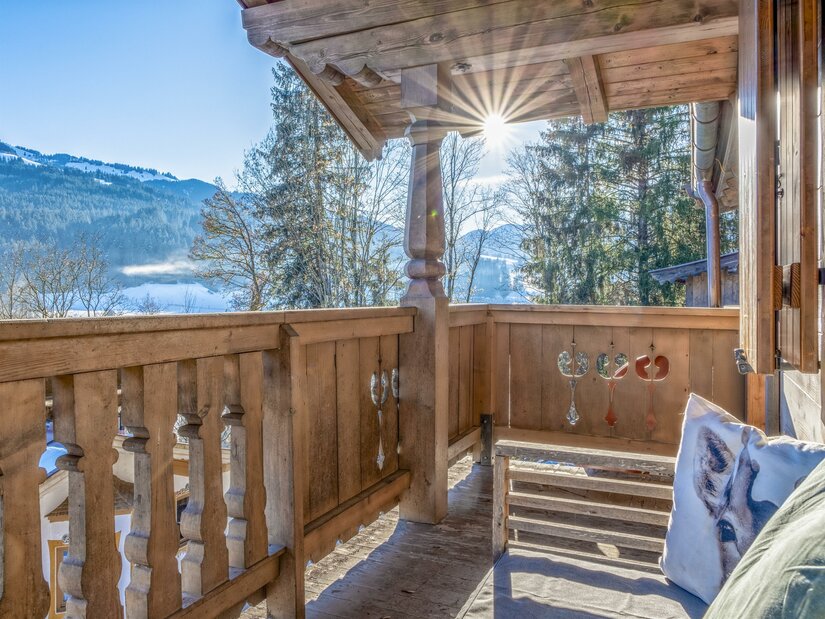Holzbalkon eines Chalets mit Blick auf verschneite Berge und Wälder bei Sonnenschein.