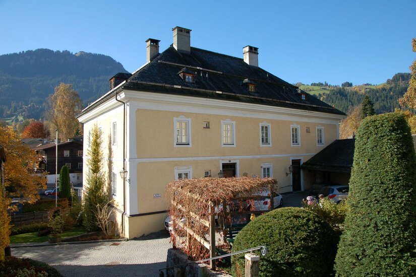 Stadtarchiv Kitzbühel Ein historisches Haus mit gelber Fassade und einem schönen Garten. Im Hintergrund sind Berge und ein blauer Himmel sichtbar.