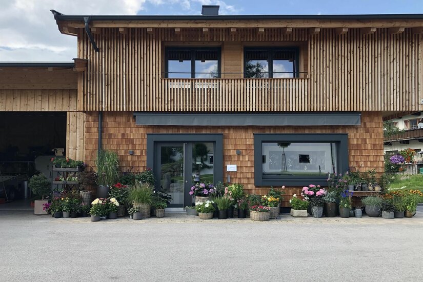Blütenwerkstatt Modernes Holzgebäude mit Schindelfassade und zahlreichen Blumenkübeln vor dem Eingang.