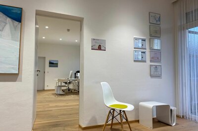 Heller Warteraum mit weißem Stuhl, Parkettboden und Blick in ein angrenzendes Büro.