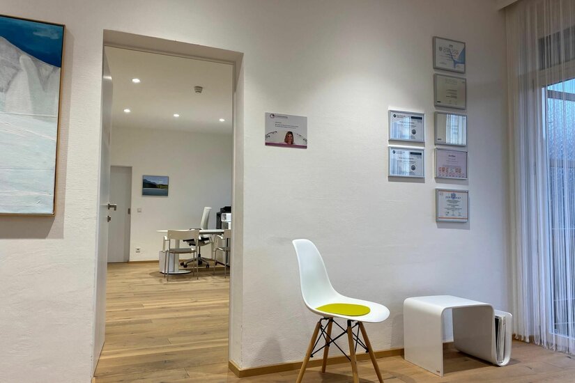 Wartebereich der Praxis Heller Warteraum mit weißem Stuhl, Parkettboden und Blick in ein angrenzendes Büro.