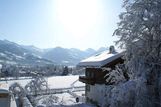 Haus Eugenie in Kitzbühel Verschneites Holzhaus vor einem weiten Tal mit schneebedeckten Bergen im Hintergrund bei Sonnenschein.