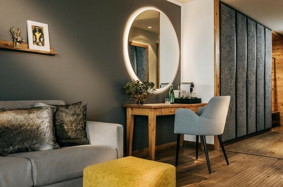 Zimmer im Grand Tirolia Modernes Hotelzimmer mit Sofa, Holztisch, beleuchtetem Rundspiegel und grauem Sessel.
