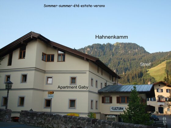 Mehrstöckiges helles Gebäude mit Satteldach vor bewaldetem Berghang unter blauem Himmel.
