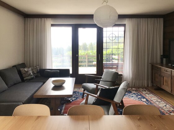 Wohnzimmer mit grauem Ecksofa, Holztisch, zwei Sesseln und großem Fenster zur Terrasse.