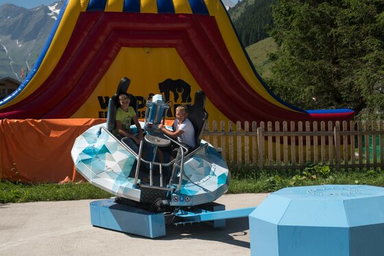 Zwei Kinder sitzen in einem rotierenden Fahrgeschäft vor einer Hüpfburg im Wildpark Ferleiten.