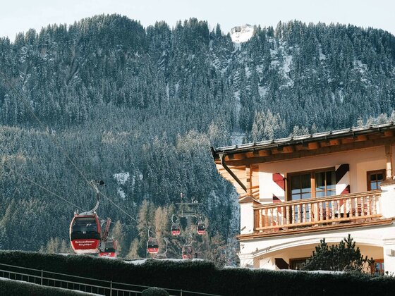Rote Gondeln der Hahnenkammbahn fahren vor einem bewaldeten Berghang an einem Hotelgebäude vorbei.