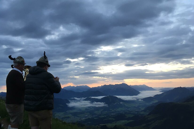 Horngipfel Zwei Männer stehen auf einem Berg und betrachten die Landschaft. Der Himmel ist bewölkt, und die Sonne geht am Horizont unter.