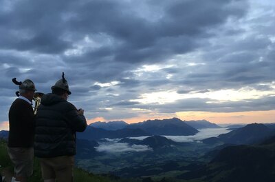 Zwei Männer stehen auf einem Berg und betrachten die Landschaft. Der Himmel ist bewölkt, und die Sonne geht am Horizont unter.