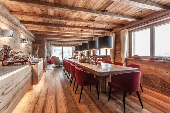 Hahnenkamm Lodge Speisebereich Rustikaler Speisesaal mit langem Holztisch, roten Polsterstühlen und Deckenbalken aus Altholz.