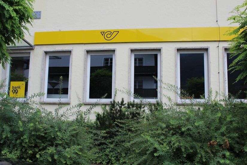 Postamt Kitzbühel Außenansicht eines Postamtes mit gelbem Logo über den Fenstern und grünen Sträuchern im Vordergrund.