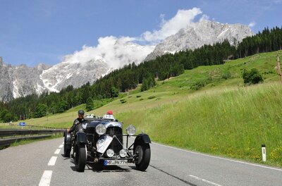 Ein schwarzer Oldtimer fährt auf einer asphaltierten Bergstraße vor einer alpinen Bergkulisse mit Wald.