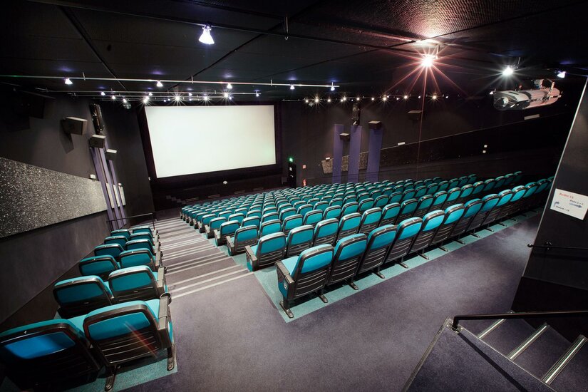 Ein modernes Kino mit blauen Sitzen und einem großen Bildschirm. Der Raum ist bereit für die nächste Filmvorführung.