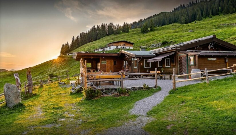Auracher Hochwildalm im Sommer Holzhütte auf einer grünen Almwiese vor bewaldetem Hang bei Sonnenuntergang.