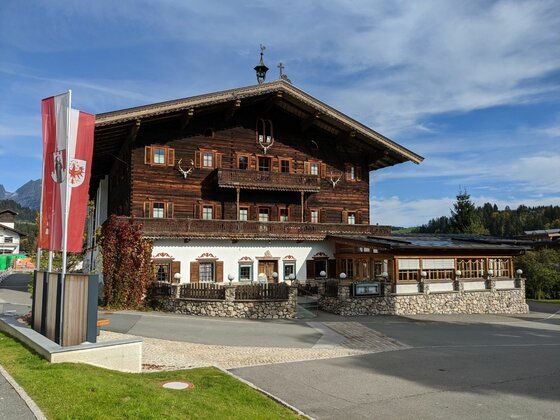 Traditionelles Alpenhaus aus dunklem Holz mit weißem Erdgeschoss und rot-weißen Fahnen davor.