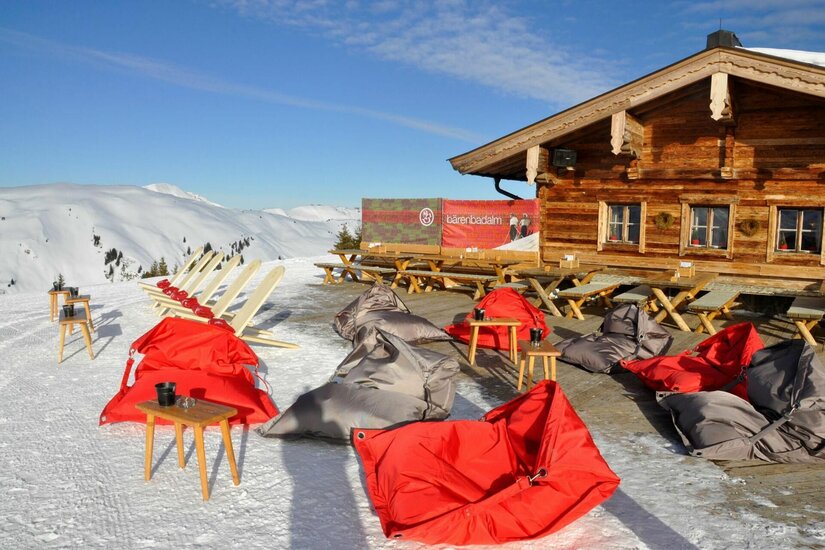 Terrasse der Bärenbadalm Sonnenterrasse der Bärenbadalm mit roten und grauen Sitzsäcken im Schnee vor einer Holzhütte.