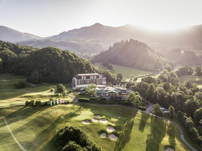 Aussenansicht Grand Tirolia Kitzbühel Luftaufnahme des Hotels Grand Tirolia Kitzbühel inmitten eines Golfplatzes vor bewaldeten Bergen bei Gegenlicht.