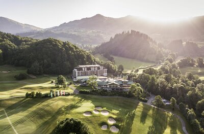 Luftaufnahme des Hotels Grand Tirolia Kitzbühel inmitten eines Golfplatzes vor bewaldeten Bergen bei Gegenlicht.