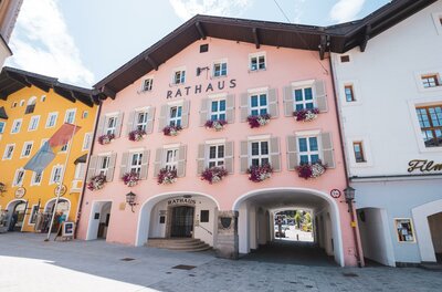Rathaus Kitzbühel Ein farbenfrohes Rathaus in einer malerischen Straße. Die Fassade ist rosa mit Blumenfensterbänken und umgeben von weiteren schönen Gebäuden.